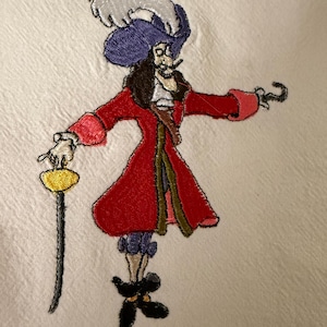 May include: An embroidered design on white fabric featuring a pirate character wearing a red coat, purple hat with a white feather, and dark breeches. The figure holds a yellow-hilted sword in one hand and has a hook on the other arm.