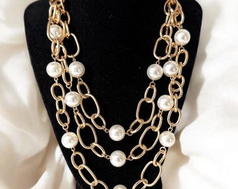 May include: A three-strand necklace with gold-colored chain links and large, round, white faux pearls. The necklace is displayed on a black jewelry bust against a white fabric backdrop.