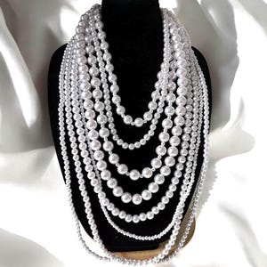 Peut inclure: Un collier de perles à plusieurs rangs exposé sur un mannequin noir. Le collier présente des perles blanches de différentes tailles, créant un effet superposé. Le fond est un tissu blanc drapé.