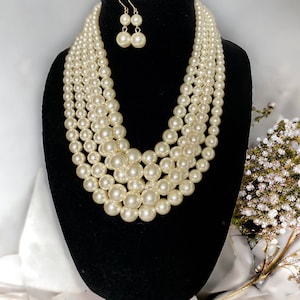 May include: A cream-colored, multi-strand pearl necklace and matching earrings set. The necklace features graduated sizes of round pearls, creating a layered effect. The earrings are small, dangling pearl drops. The jewelry is displayed on a black mannequin.