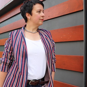 May include: A blazer featuring a vertical striped pattern in navy blue, red, and white, worn over a plain white top. The blazer has long sleeves and a shawl collar. A silver-toned beaded necklace is worn around the neck.
