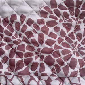 Könnte beinhalten: Ein weißer gesteppter Stoff mit einem sich wiederholenden Muster aus burgunderfarbenen Blumendesigns. Der Stoff hat den Text "Laura Louisse" in die obere rechte Ecke gestickt.