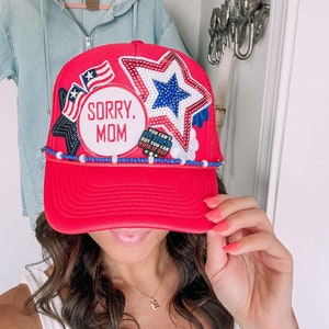 May include: A red trucker hat with a curved peak. The hat has patriotic patches, including stars, stripes, and the text "SORRY, MOM." Additional text reads "YOU CAN'T FIX STUPID BUT YOU CAN VOTE IT OUT." The hat is decorated with blue and white beads.