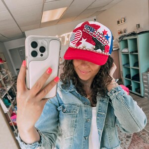 May include: A red, white, and blue baseball cap with the word "TRUMP" in red and blue letters. The cap features stars, hearts, and a chain around the brim. The person in the image is wearing a denim jacket.
