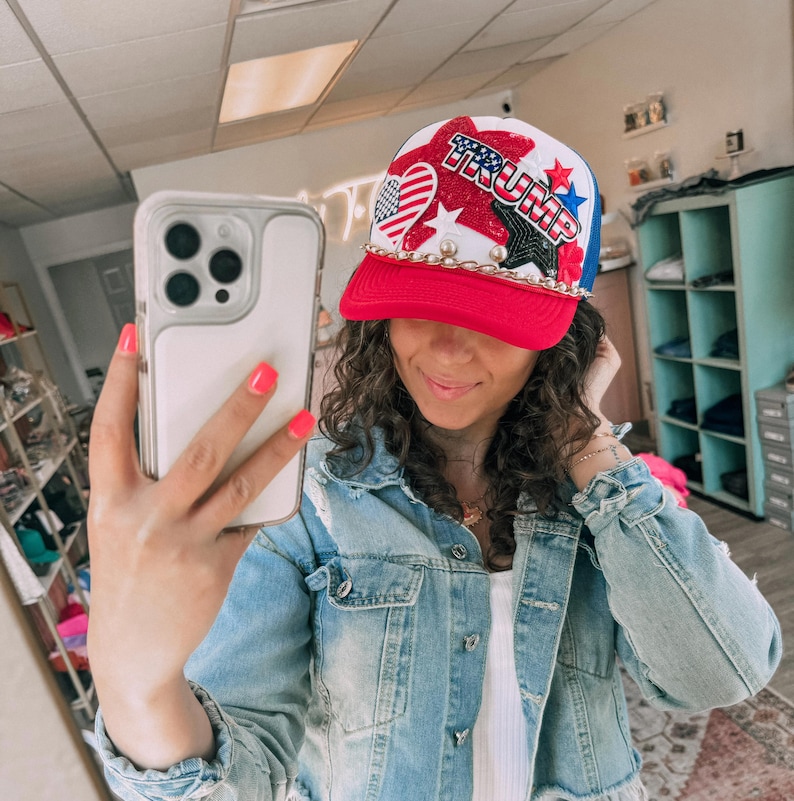 May include: A red, white, and blue baseball cap with the word "TRUMP" in red and blue letters. The cap features stars, hearts, and a chain around the brim. The person in the image is wearing a denim jacket.