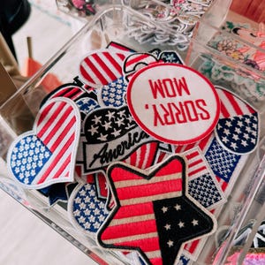 May include: A clear container filled with assorted patriotic patches. The patches feature red, white, and blue designs, including stars, stripes, and the word "America." One patch reads "Sorry Mom, Wow."