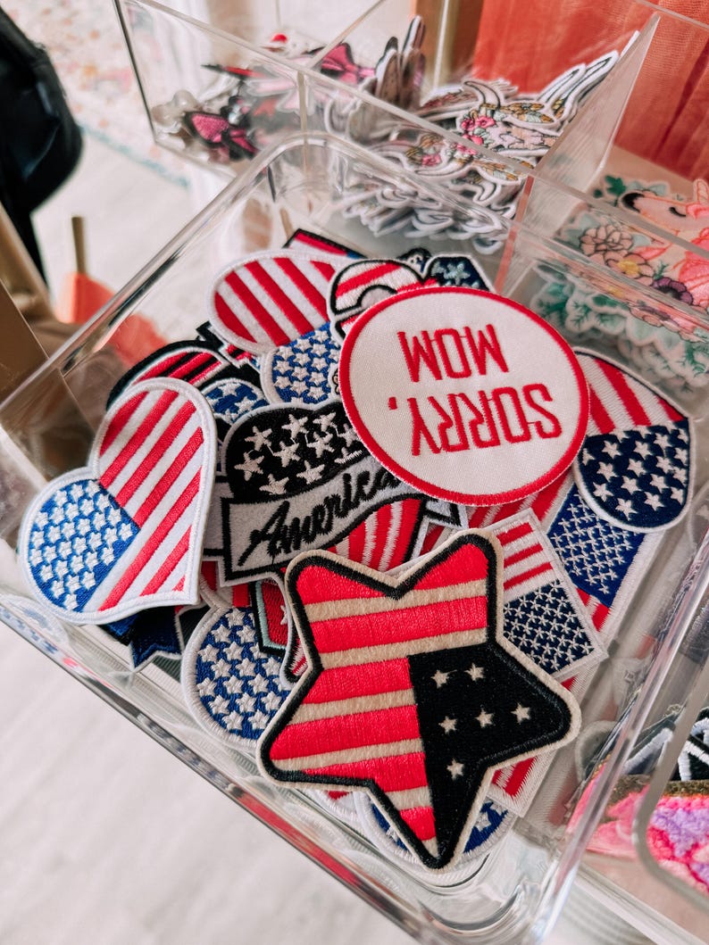 May include: A clear container filled with assorted patriotic patches. The patches feature red, white, and blue designs, including stars, stripes, and the word "America." One patch reads "Sorry Mom, Wow."