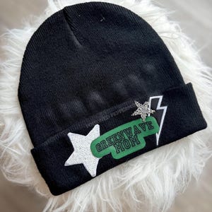 May include: Black beanie with a white star and a green rectangle with the text "GREENWAVE MOM". A silver glitter star and white lightning bolt are also on the beanie. The beanie is on a white fluffy surface.