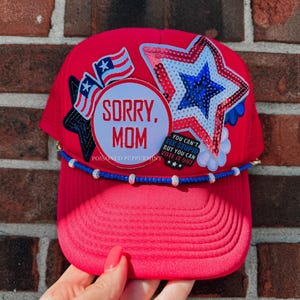 May include: A red trucker hat with a curved peak, adorned with patriotic patches and the text "SORRY, MOM." It features a sequined star, American flags, and a beaded band. The hat has a mesh back.