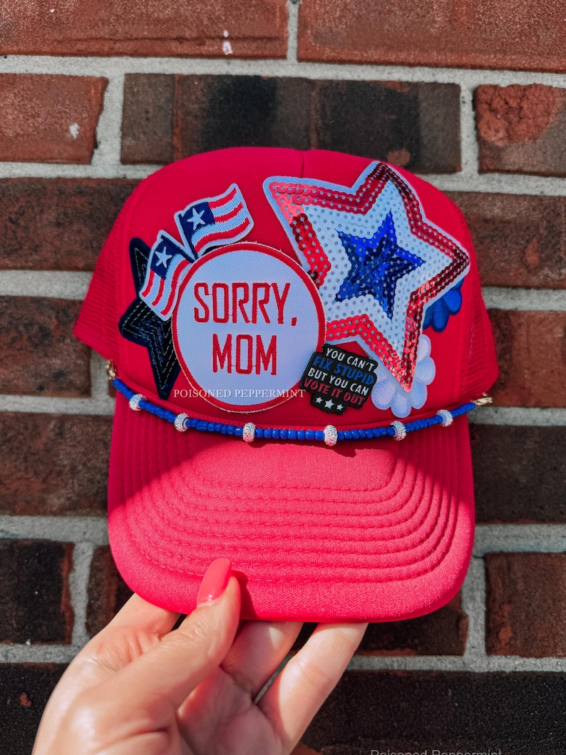 May include: A red trucker hat with a curved peak, adorned with patriotic patches and the text "SORRY, MOM." It features a sequined star, American flags, and a beaded band. The hat has a mesh back.