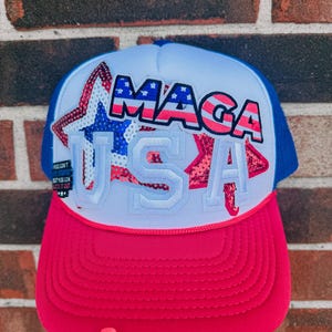 May include: A red, white, and blue trucker hat with the text "MAGA USA" in a patriotic design. The hat features a red brim, a white front panel with a star graphic, a blue mesh back, and a curved brim.