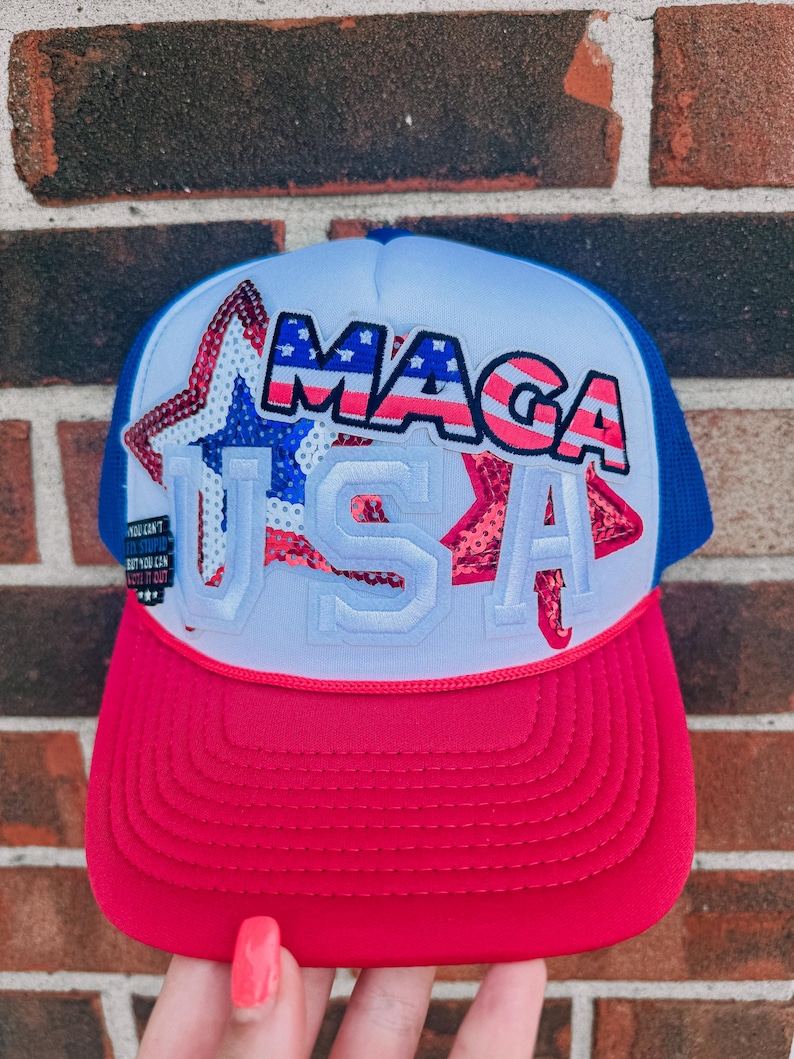 May include: A red, white, and blue trucker hat with the text "MAGA USA" in a patriotic design. The hat features a red brim, a white front panel with a star graphic, a blue mesh back, and a curved brim.