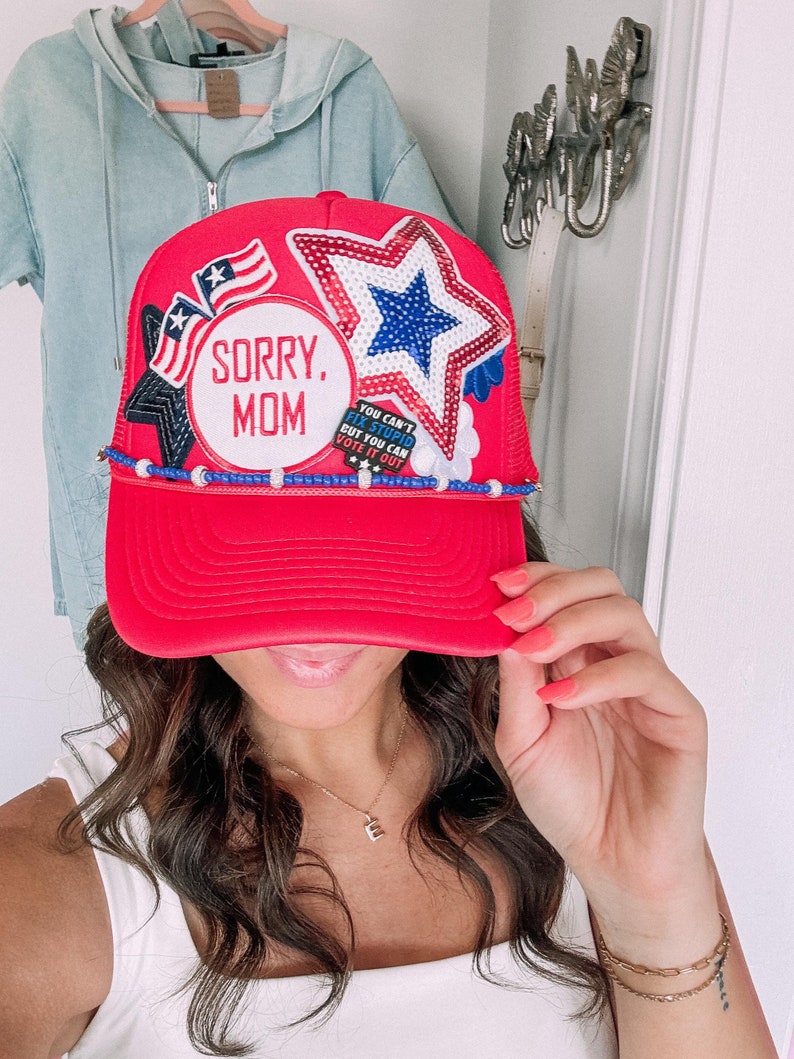May include: A red trucker hat with a curved peak. The hat has patriotic patches, including stars, stripes, and the text "SORRY, MOM." Additional text reads "YOU CAN'T FIX STUPID BUT YOU CAN VOTE IT OUT." The hat is decorated with blue and white beads.