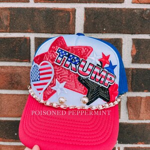May include: A red and blue trucker hat with the word "TRUMP" in white, red, and blue. The hat features a white front panel with sequined stars and a heart with an American flag design. A gold chain and the text "POISONED PEPPERMINT" are also visible.