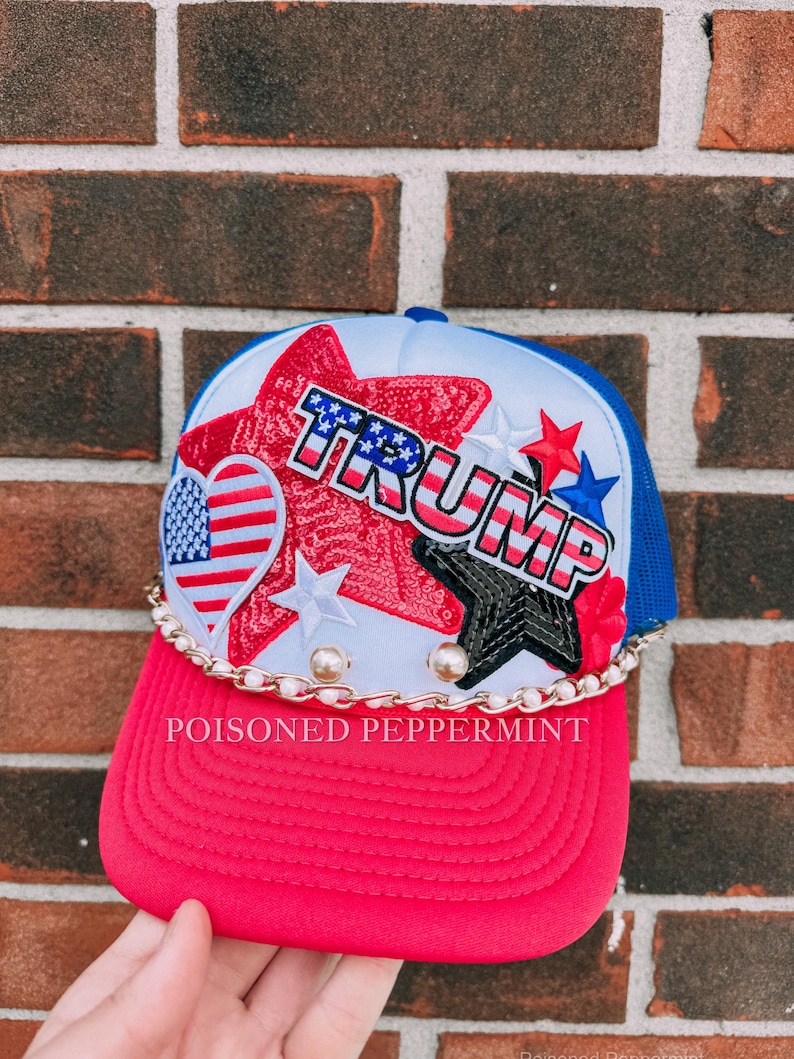 May include: A red and blue trucker hat with the word "TRUMP" in white, red, and blue. The hat features a white front panel with sequined stars and a heart with an American flag design. A gold chain and the text "POISONED PEPPERMINT" are also visible.
