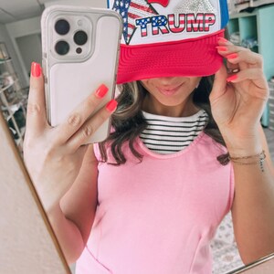 May include: A red, white, and blue trucker hat with the word "TRUMP" across the front. The hat features a sequined American flag and star design. The hat has a red brim and a white front panel with a blue mesh back.