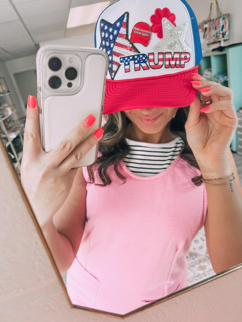 May include: A red, white, and blue trucker hat with the word "TRUMP" across the front. The hat features a sequined American flag and star design. The hat has a red brim and a white front panel with a blue mesh back.