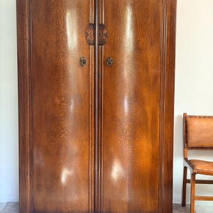 May include: A tall, two-door wooden wardrobe with a polished medium-brown finish and visible wood grain. The doors feature arched tops, decorative carved wooden plates near the centre, and circular metal pull handles with a keyhole below.