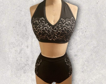Puede incluir: Un traje de baño de dos piezas negro que incluye un top estilo halter con escote en V profundo y una braga de talle alto. Ambas piezas presentan un detallado patrón de encaje floral blanco sobre el busto y en los laterales de las caderas.