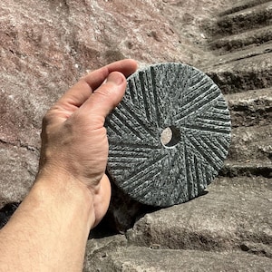 May include: A circular dark gray stone millstone featuring a central hole and a radial pattern of carved grooves across its flat surface. The stone has a speckled texture and is held in a position that displays its textured face and circular shape.