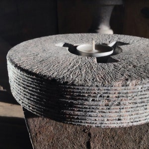 May include: A round, gray stone candle holder with a circular indentation in the center for a tea light. The holder has a textured surface and is made of layered stone.