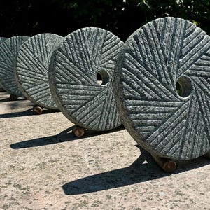 May include: Five circular gray stone millstones arranged in a row, each featuring a central hole and a radial pattern of carved grooves. The stones are supported by small wooden stands, displaying a textured, weathered surface and natural gray tones.