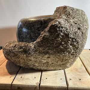 Pode incluir: Uma grande tigela de pedra esculpida à mão com um interior escuro e salpicado. A tigela é embalada por um exterior áspero e texturizado, criando uma estética natural e orgânica. A peça repousa sobre uma superfície de madeira.