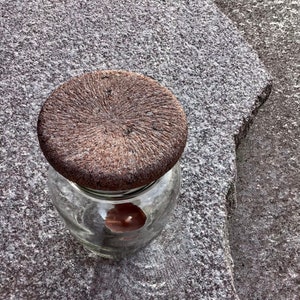 Könnte beinhalten: Ein braunes Glasgefäß mit einem strukturierten Deckel. Das Gefäß ist durchsichtig und zeigt eine braune Kerze im Inneren.