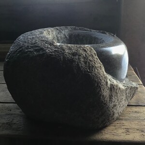 Può includere: Una grande ciotola rotonda in pietra di colore grigio scuro con un interno liscio e lucido. La ciotola è realizzata in pietra ruvida e testurizzata e ha un bordo leggermente irregolare.