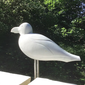 Peut inclure: Une sculpture d'oiseau en marbre blanc, posée sur un support en métal. La sculpture est dans un jardin avec de la végétation verte en arrière-plan.