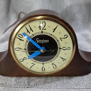 May include: A vintage Sessions clock with a dark wood case and a gold-tone bezel. The clock face is cream-colored with black numerals and blue hands. The Sessions logo is displayed in the center.