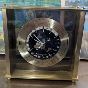 May include: A gold-toned Seiko world clock with a glass enclosure. The clock face features a black background with a world map and city names. The clock hands are silver, and the clock displays the time and date.