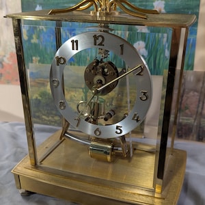 May include: A gold-toned, rectangular clock with a clear glass enclosure. The clock face is silver with black numerals. The clock has a pendulum and decorative gold accents on top. The clock sits on a gold base.