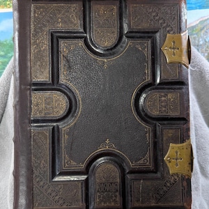 May include: A vintage, dark brown leather-bound book with ornate gold-colored embossed designs. The book features a central cross-shaped design and decorative corner clasps. The spine is a darker shade of brown.