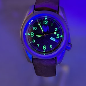 May include: A black watch with a silver case and a brown leather band. The watch face has luminous green hands and markers, and the day of the week is displayed as "WED".