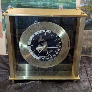 May include: A square Seiko quartz world time clock with a gold-tone metal frame and a glass front. The circular dial features a black center with a world map graphic, surrounded by a gold ring displaying 24-hour markers and global city names.