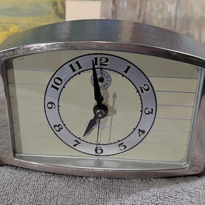 May include: A vintage silver-toned alarm clock with a cream-colored face. The clock features black numerals and hands, with a smaller dial at the top. The clock has a rectangular shape with a curved top and side supports.