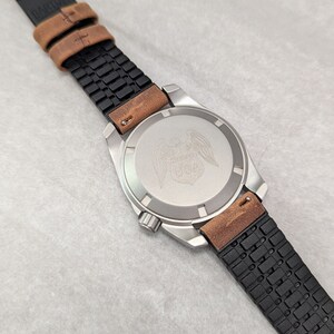 May include: A silver watch with a black and brown leather strap. The watch face has a silver eagle design with wings spread out. The strap has a textured pattern.