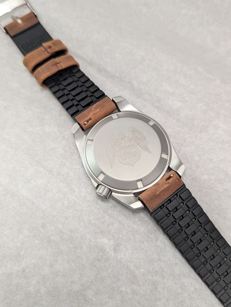 May include: A silver watch with a black and brown leather strap. The watch face has a silver eagle design with wings spread out. The strap has a textured pattern.