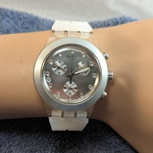 May include: A Swatch wristwatch with a silver dial and white band. The watch face has three sub-dials and a date display. The watch is on a wrist.