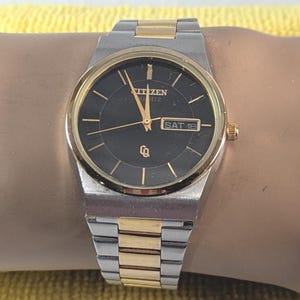 May include: A Citizen Quartz watch with a black dial, gold-tone bezel, and silver and gold-tone band. The watch face displays the day and date. The watch is a classic design with a rectangular case and a link bracelet.