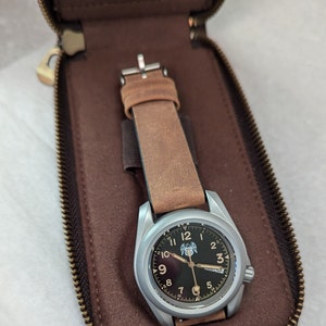 May include: A silver wristwatch with a brown leather strap in a brown leather case. The watch face has black numerals and a black background with a silver eagle logo.