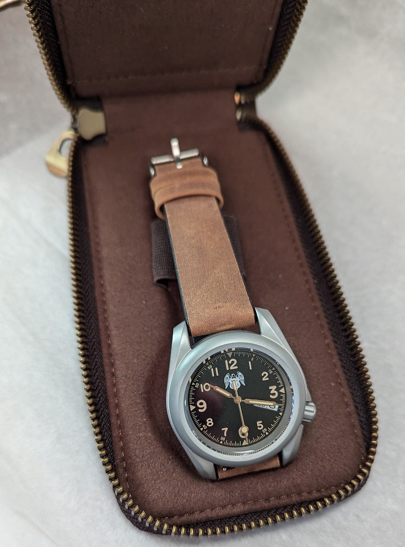 May include: A silver wristwatch with a brown leather strap in a brown leather case. The watch face has black numerals and a black background with a silver eagle logo.