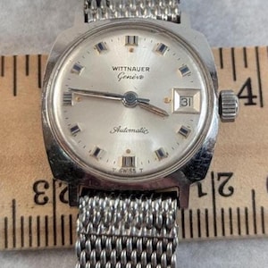 May include: Vintage Wittnauer Genève automatic wristwatch with a silver-tone dial and mesh band. The watch face displays the date and the words "Wittnauer Genève Automatic". The watch has a silver-tone case and a mesh metal band.