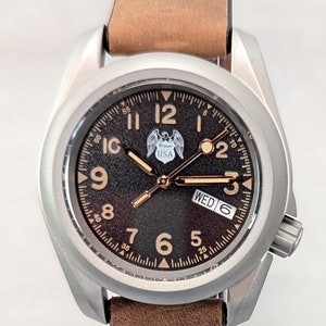 May include: A silver-toned wristwatch with a brown leather band. The watch face is black with white markings and a small eagle logo with the word "USA". The day of the week is displayed as "WED" and the date is "6".