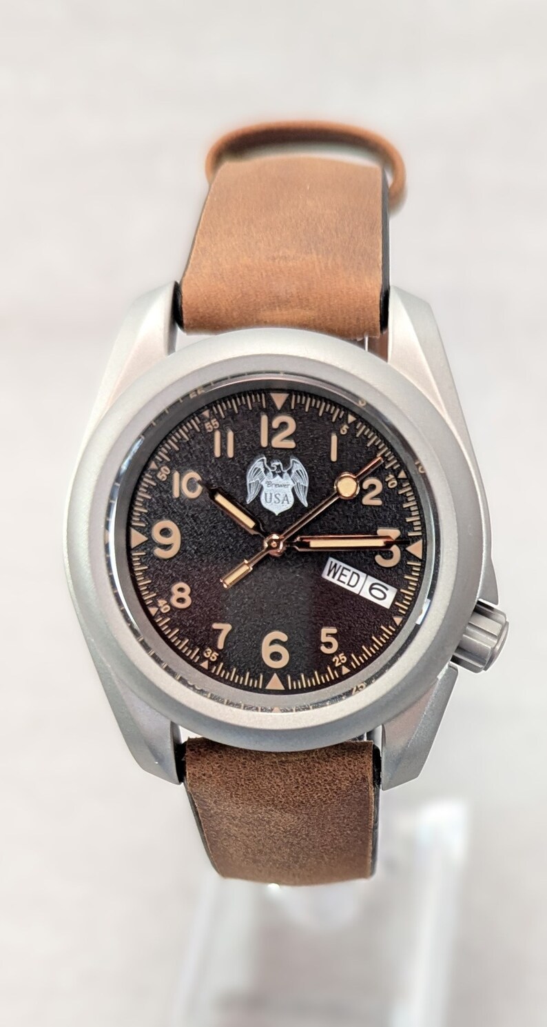 May include: A silver-toned wristwatch with a brown leather band. The watch face is black with white markings and a small eagle logo with the word "USA". The day of the week is displayed as "WED" and the date is "6".