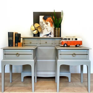 Op de afbeelding: Een grijze slaapkamer set met twee nachtkastjes en een dressoir. Elk nachtkastje heeft een lade met een goudkleurige ringgreep. Een speelgoed oranje en witte VW bus staat bovenop het dressoir. Een ingelijste foto en boeken zijn ook te zien.