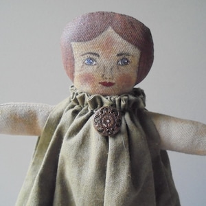 K&ouml;nnte beinhalten: Eine handgemachte Stoffpuppe mit bemaltem Gesicht, braunen Haaren und einem gr&uuml;nen Kleid. Die Puppe hat einen Knopf auf der Vorderseite des Kleides.