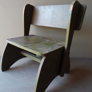 May include: A vintage wooden chair painted in shades of green and gray. The chair features a backrest and a seat, with curved legs and supports. The paint shows signs of wear, giving it a rustic appearance. The chair's dimensions are approximately 24 inches high, 12 inches wide, and 12 inches deep.