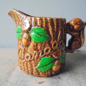 May include: A brown ceramic creamer pitcher shaped like a tree stump with a squirrel handle. The pitcher has green leaves and acorns painted on it and the word "Canada" written on the side.
